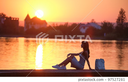 Lonely woman sitting alone on lake shore on warm evening. Solitude and relaxing in nature concept. 97527281