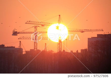 Dark silhouette of tower cranes with big setting sun at high residential apartment buildings construction site at sunset. Real estate development 97527806