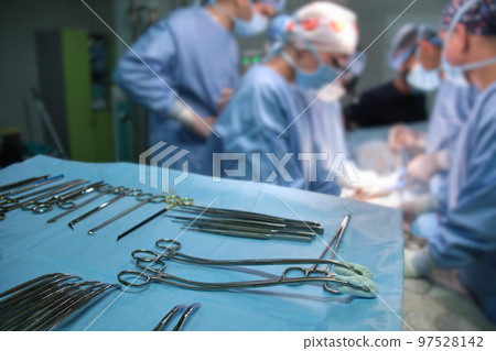 Closeup of surgical tools on table and team of surgery doctors operating a patient in hospital room Closeup of surgical tools on table and team of surgery doctors operating a patient in hospital room 97528142