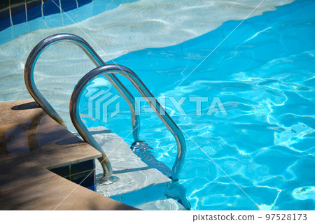 Close up of swimming pool stainless steel handrail descending into tortoise clear pool water. Accessibility of recreational activities concept. Close up of swimming pool stainless steel handrail descending into tortoise clear pool water. Accessibility of recreational activities concept. 97528173