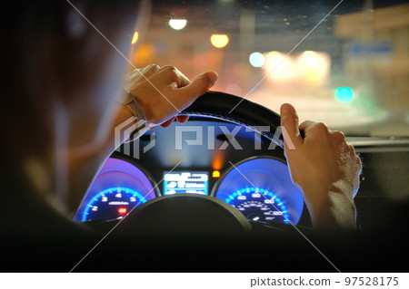 Close up of driver hands holding steering wheel driving car with blurred city street lights on background at night Close up of driver hands holding steering wheel driving car with blurred city street lights on background at night 97528175