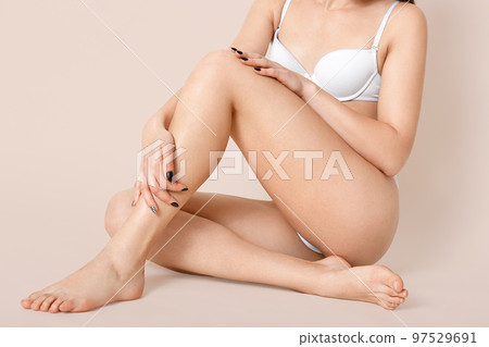 Cropped shot of half naked young woman wears white lingerie, shows slender legs, has healthy smooth skin, crossed legs, sits on floor against beige background, has perfect figure, poses bare foot. Cropped shot of half naked young woman wears white lingerie, shows slender legs, has healthy smooth skin, crossed legs, sits on floor against beige background, has perfect figure, poses bare foot. 97529691