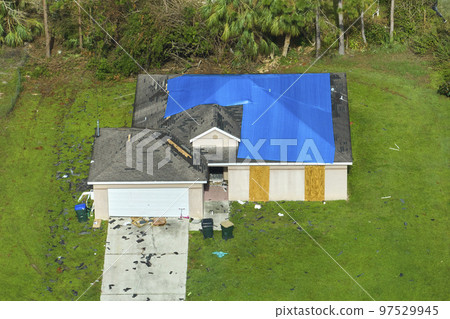Aerial view of damaged in hurricane Ian house roof covered with blue protective tarp against rain water leaking until replacement of asphalt shingles 97529945