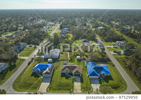 Aerial view of damaged in hurricane Ian house roof covered with blue protective tarp against rain water leaking until replacement of asphalt shingles 97529950