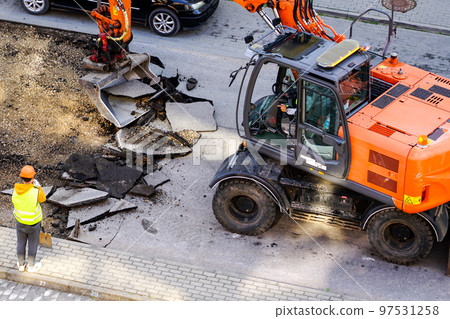 An excavator breaks up the old asphalt layer with a bucket during street repairs 97531258