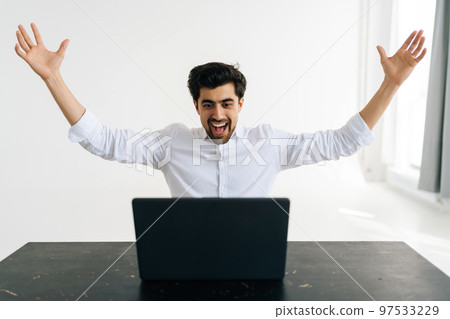 Studio portrait of cheerful excited male office worker screaming eureka raising hands up, solving hard task, getting access, successfully completing work sitting at desk in room with light interior. Studio portrait of cheerful excited male office worker screaming eureka raising hands up, solving hard task, getting access, successfully completing work sitting at desk in room with light interior. 97533229