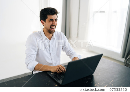 Studio portrait of happy excited young man in shirt using working on laptop computer smiling looking away sitting at table in room with light interior. Cheerful male freelancer at remote home office. Studio portrait of happy excited young man in shirt using working on laptop computer smiling looking away sitting at table in room with light interior. Cheerful male freelancer at remote home office. 97533230