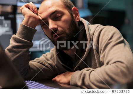 Tired male programmer falling asleep at office desk working on interface programming, holding head with hand late at night. Exhausted overworked developer sleeping after coding database system. 97533735