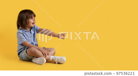 Cute little boy pointing aside at empty space, sitting on floor and enjoying what he sees, orange background, panorama Cute little boy pointing aside at empty space, sitting on floor and enjoying what he sees, orange background, panorama 97536221