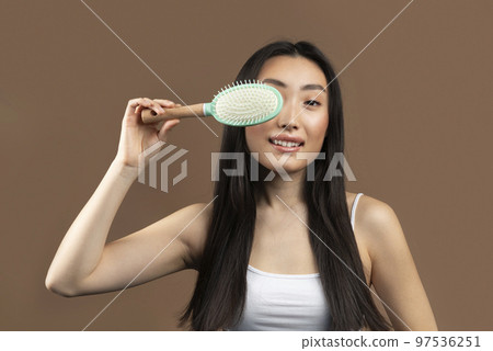 Hair care concept. Excited korean woman with long hair covering eye with hair brush and smiling over brown background 97536251