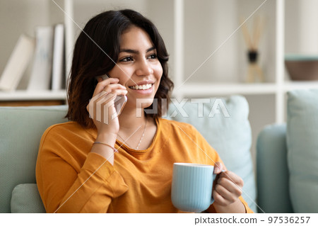 Smiling Young Arab Woman Talking On Cellphone And Drinking Coffee At Home Smiling Young Arab Woman Talking On Cellphone And Drinking Coffee At Home 97536257