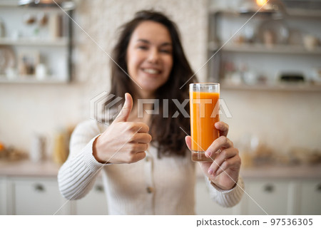 Positive caucasian lady holding glass of organic orange juice and showing thumb up, standing in light kitchen interior 97536305