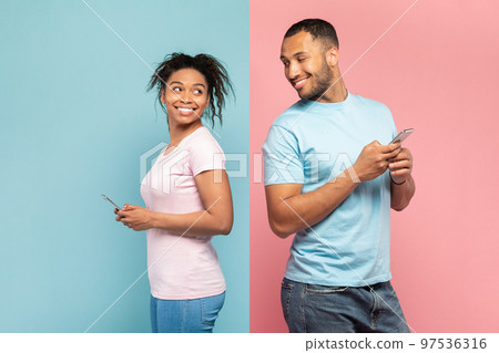 Curious black man using smartphone, looking and peeking at his girlfriend's phone, standing on pink and blue background Curious black man using smartphone, looking and peeking at his girlfriend's phone, standing on pink and blue background 97536316