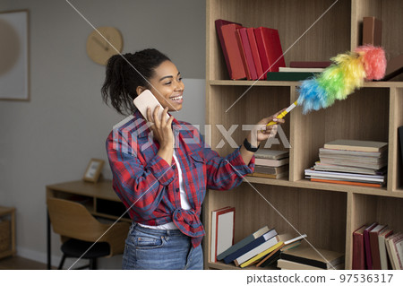 Smiling millennial african american lady dusting and calling by phone, enjoy household chores in living room Smiling millennial african american lady dusting and calling by phone, enjoy household chores in living room 97536317
