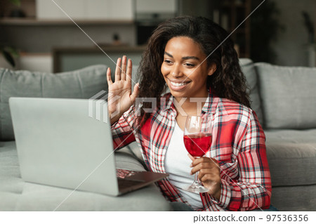 Glad millennial black lady waving hand, say hello greeting in computer camera, hold glass of wine in living room Glad millennial black lady waving hand, say hello greeting in computer camera, hold glass of wine in living room 97536356
