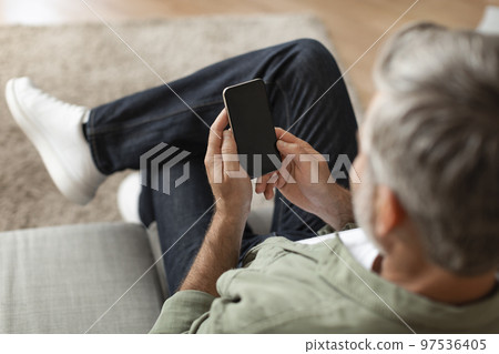 Elderly caucasian man typing on smartphone with empty screen using new app for chat, sit on sofa 97536405