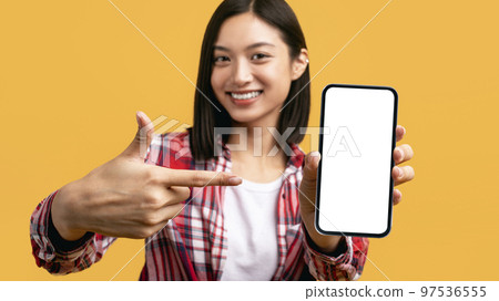 Mobile app. Happy asian lady pointing at cellphone with blank screen over yellow background, panorama, mockup 97536555