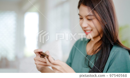 Happy young asian woman relax on comfortable couch at home texting messaging on smartphone Happy young asian woman relax on comfortable couch at home texting messaging on smartphone 97540568