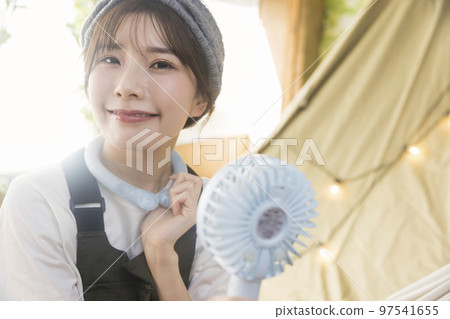 A young woman who cools down with a neck cooler and a portable fan at a house camp Camping image 97541655