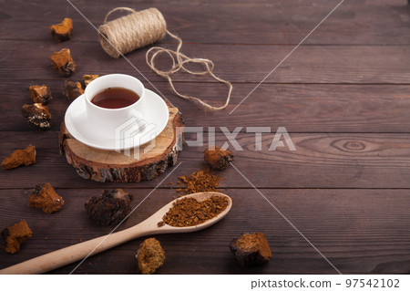 Chaga mushroom coffee drink in a white cup on a wooden stand, scattered pieces of chaga and a spoon with chaga powder on a wooden background. Copy spaes. 97542102
