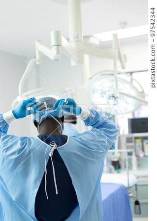 Rear of african american surgeon in gown and gloves tying mask in theatre, vertical with copy space 97542344