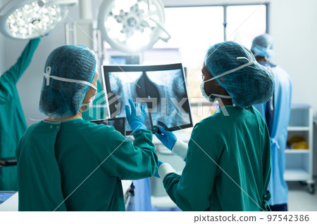 Rear view of two diverse female surgeons discussing x-ray in operating theatre, with colleagues 97542386