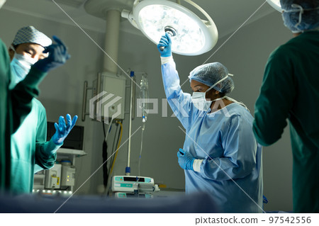 Biracial female surgical tech adjusting light in operating theatre during operation 97542556