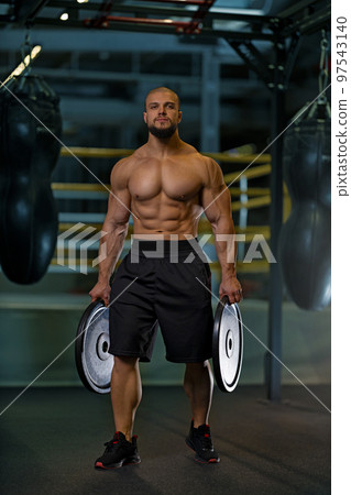 Athletic man with naked torso holding black plates in the gym Athletic man with naked torso holding black plates in the gym 97543140