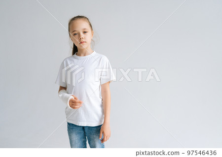 Portrait of sad blonde little girl with broken hand wrapped in white plaster bandage standing on white isolated background in studio, looking at camera. Concept of child insurance and healthcare. 97546436
