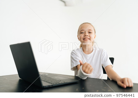 Happy blonde little girl showing thumb-up gesture with broken hand wrapped in white plaster bandage sitting at table with laptop smiling looking at camera. Concept of child insurance and healthcare. 97546441