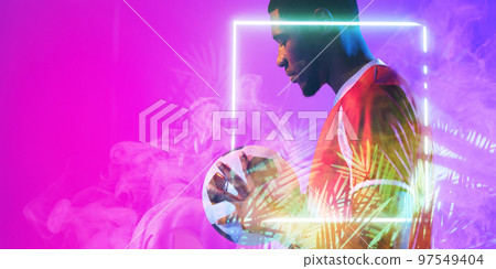 African american male soccer player with ball standing by illuminated plant and square African american male soccer player with ball standing by illuminated plant and square 97549404