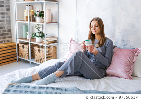 Attractive brunette woman is sitting on bed in her pajamas drinking hot tea or 97550617