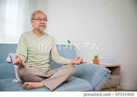 Lifestyle calm senior man lotus pose doing yoga for mental balance breathing air relaxing on sofa at home, Asian elderly old man sitting practise yoga and meditation in lotus position and closed eyes 97553409