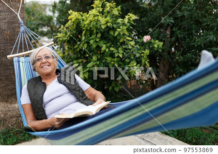 Smiling senior good-looking grey hair woman wearing glasses while reading in hammock in the summer garden 97553869