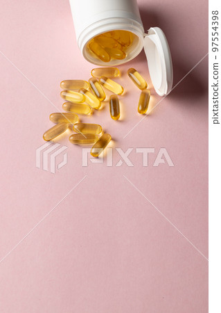 Vertical composition of white pill box spilling oil capsules on pink background with copy space Vertical composition of white pill box spilling oil capsules on pink background with copy space 97554398