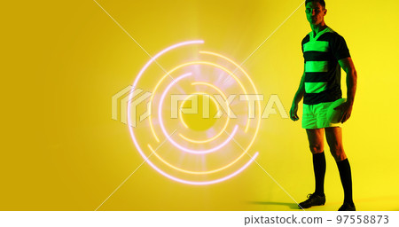 Full length of caucasian rugby player with ball standing by illuminated circular pattern, copy space 97558873