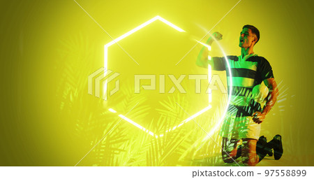Caucasian rugby player with ball screaming and shaking fist by illuminated hexagon and plants 97558899