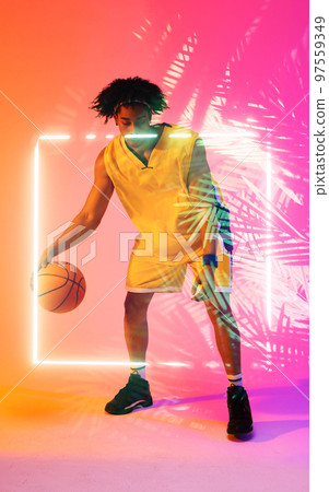 Full length of biracial male player dribbling basketball by illuminated plants and square 97559349