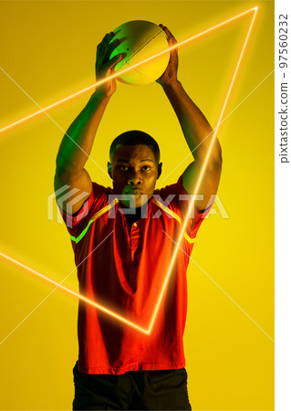 African american male rugby player throwing ball over illuminated triangle on yellow background 97560232