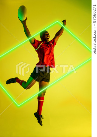 African american male player with rugby ball raising arms and jumping by illuminated rectangle 97560271