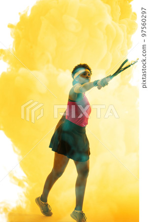 Full length of african american female player hitting with racket over yellow smoky background 97560297