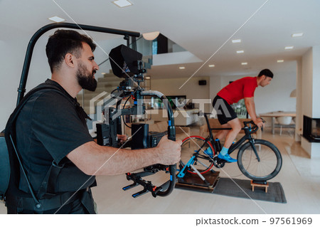 A cameraman filming an athlete riding a triathlon bike on a simulation machine in a modern living room. Training in pandemic conditions. A cameraman filming an athlete riding a triathlon bike on a simulation machine in a modern living room. Training in pandemic conditions. 97561969