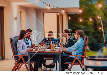 A group of young diverse people having dinner on the terrace of a modern house in the evening. Fun for friends and family. Celebration of holidays, weddings with barbecue. 97562103