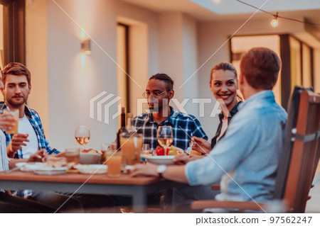 A group of young diverse people having dinner on the terrace of a modern house in the evening. Fun for friends and family. Celebration of holidays, weddings with barbecue. 97562247