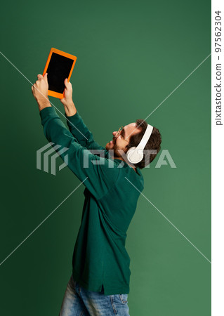 Portrait of young handsome happy man with beard wearing casual sweater and glasses showing tablet over green background. Happiness, positive emotions Portrait of young handsome happy man with beard wearing casual sweater and glasses showing tablet over green background. Happiness, positive emotions 97562304