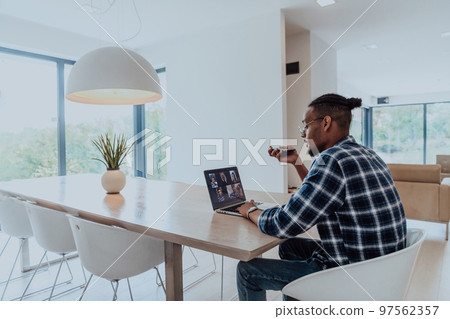 African American man in glasses sitting at a table in a modern living room, using a laptop and smartphone for business video chat, conversation with friends and entertainment African American man in glasses sitting at a table in a modern living room, using a laptop and smartphone for business video chat, conversation with friends and entertainment 97562357
