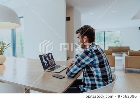 The man sitting at a table in a modern living room, with headphones using a laptop for business video chat, conversation with friends and entertainment 97562390