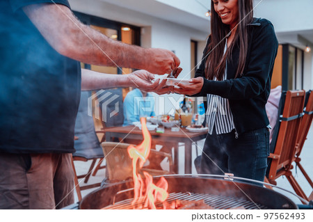 A group of friends and family barbecue together in the evening on the terrace in front of a large modern house 97562493