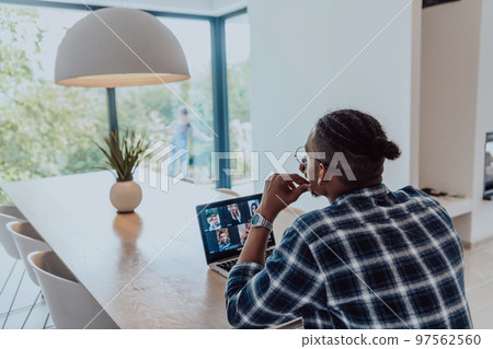 African American man in glasses sitting at a table in a modern living room, using a laptop and smartphone for business video chat, conversation with friends and entertainment African American man in glasses sitting at a table in a modern living room, using a laptop and smartphone for business video chat, conversation with friends and entertainment 97562560