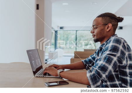 African American man in glasses sitting at a table in a modern living room, using a laptop for business video chat, conversation with friends and entertainment African American man in glasses sitting at a table in a modern living room, using a laptop for business video chat, conversation with friends and entertainment 97562592